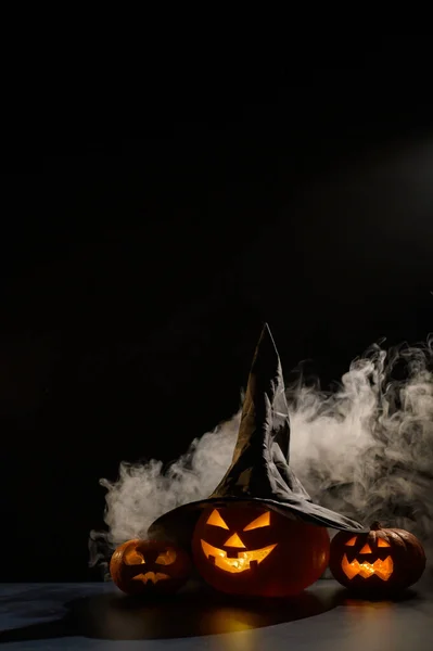 Biglietto di Halloween verticale. Cappello da strega su una zucca con smorfie raccapriccianti scolpite su fondo nero nella nebbia. Jack-o-lanterna brilla dall'interno ed emette fumo. — Foto Stock