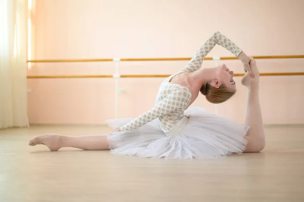 Belle ballerine dans le corps et tutu blanc s'entraîne dans un cours de danse. Jeune danseuse flexible posant dans des chaussures pointes assises sur le sol. — Photo