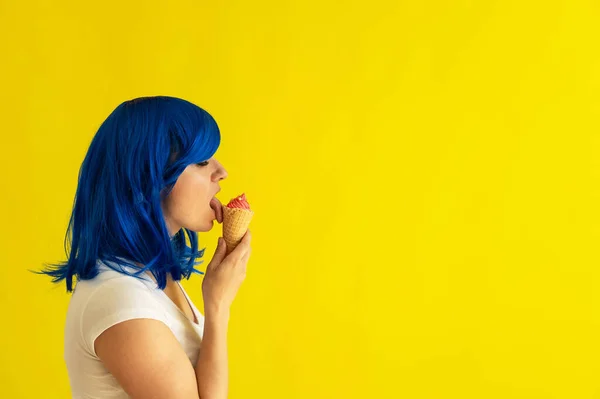 Красивая женщина с голубыми волосами стоит в профиль и облизывает рог малинового мороженого на желтом фоне. Девушка в парике любит есть мороженое в вафельной чашке. Летний охлаждающий десерт. — стоковое фото