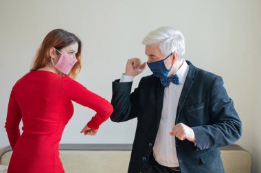 Olgun, gri saçlı bir adam ve kırmızı saçlı, maskeli bir kadın dirseklerini selamlıyor. El sıkışmaya ve sarılmaya bir alternatif. Meslektaşlar aralarındaki mesafeyi koruyorlar. Popüler bir selamlama şekli..