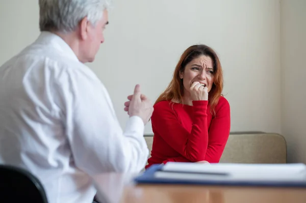 An upset woman bites her nails at a session with a male psychologist. Mature gray-haired psychotherapist talking to a female patient with depression and neurosis. Unbalanced psyche. Onychophagia.