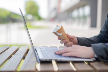 Yüzü olmayan bir kadın yazlık terasta bilgisayar başında çalışıyor ve vanilyalı dondurma yiyor. Dizüstü bilgisayarda yazı yazan ve elinde dondurma külahı tutan kadınların yakın çekimi. Yaz serinletici tatlı.