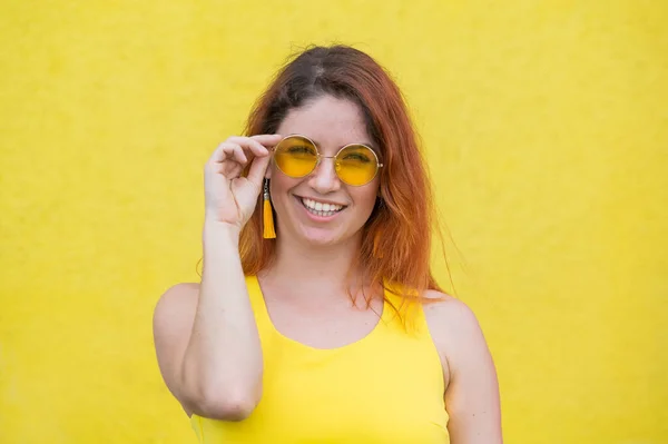 Portrét šťastné ženy ve slunečních brýlích na žlutém pozadí. Červenovlasá bezstarostná dívka v šatech a náušnicových střapcích stejné barvy. Dáma s bezvadným, oslnivým úsměvem. Letní foto. — Stock fotografie