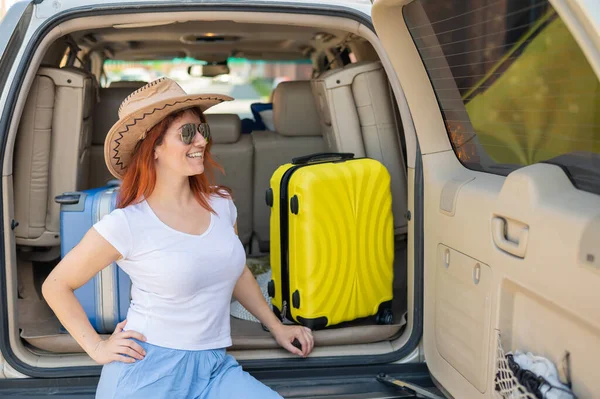 Glückliche rothaarige Frau mit Cowboyhut und Sonnenbrille auf einer Autofahrt durch das Land. Mädchen im Kofferraum mit gelbem Koffer, bereit für den Sommerurlaub. Unabhängiges Reisen. — Stockfoto