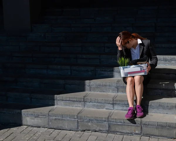 Mujer deprimida vestida con máscara médica es despedida y está sentada en las escaleras con una caja de pertenencias personales. Trabajadora de oficina con traje y zapatillas al aire libre. El desempleo en la crisis económica —  Fotos de Stock