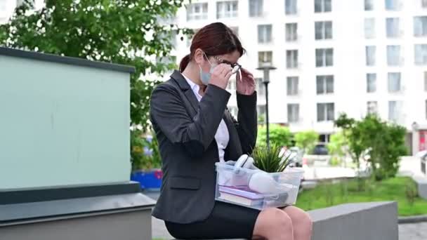 Infeliz despedido mujer enmascarada sentado en la calle con una caja de artículos personales desde el escritorio. Una mujer desesperada desempleada se quita las gafas. Reducción del empleo durante la epidemia de coronavirus . — Vídeos de Stock