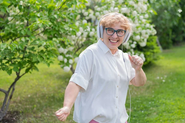 Feliz anciana escucha música en los auriculares y baila en un parque de manzanos florecientes. La activa pensionista sonríe y disfruta de un cálido día de verano en el jardín. Generación avanzada de edad. — Foto de Stock