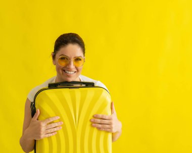 Güneş gözlüklü kar beyazı gülüşlü bir kadın sarı arka planda bir bavula sarılıyor. Heyecanlı kız yaz tatilinde seyahat için bekliyor ve çantasını sarılıyor..