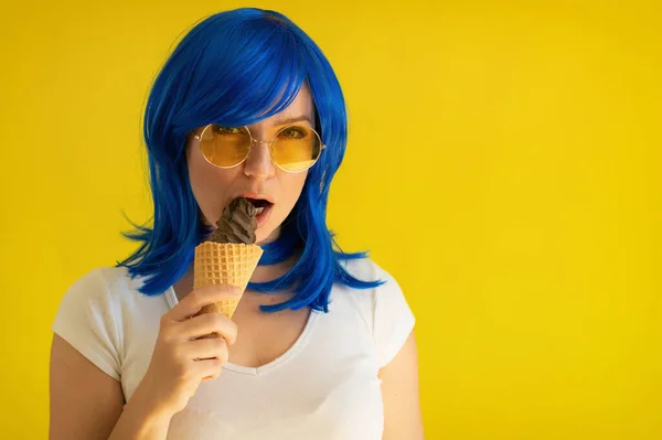 Porträtt av en vacker kvinna med blått hår gärna äta en chokladglass kon på en gul bakgrund. En flicka i solglasögon och en peruk håller en gelato i en våffelkopp. Sommar kylning dessert. — Stockfoto