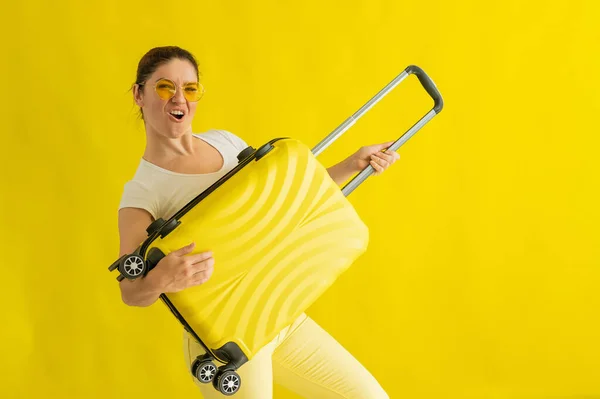 戴着太阳镜的女人鬼鬼祟祟地笑着，还拿着一只手提箱，像把吉它放在黄色的背景上。一个期待暑假旅行的兴奋女孩模拟弹奏弦乐器. — 图库照片