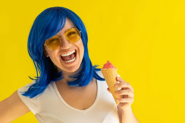 Портрет женщины с голубыми волосами в солнечных очках, держащей вафельный конус с мороженым на желтом фоне. Красивая кавказская девушка с прекрасной улыбкой наслаждается летним охлаждающим десертом. — стоковое фото