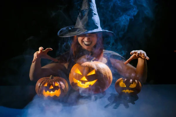 Zlověstná čarodějnice v klobouku vykouzlí nad lucernou. Tradiční halloween postavy. Tajemná mlha se plazí nad dýněmi s vyřezávanými hroznými tvářemi. Žena v karnevalovém kostýmu. — Stock fotografie