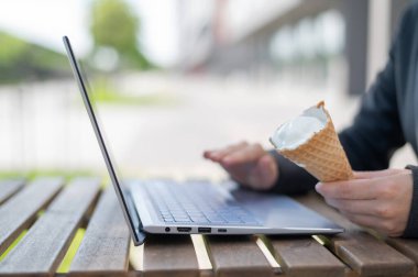 Yazlık terasta dizüstü bilgisayarlı bir klavyeyle bir iş kadınının ellerine yakın çekim. Ofis görevlisi dışarıda dondurma külahı yiyor. Kesilmiş fotoğraf. Vanilyalı dondurulmuş yoğurt..