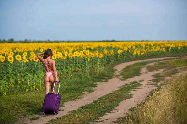 Schöne Frau im rosafarbenen Badeanzug reist mit einem Koffer durch die Landschaft. Ein Mädchen im Monokini läuft mit einer großen Tasche allein auf einem Sonnenblumenfeld. Wandern. Ruhe und soziale Distanz. — Stockfoto