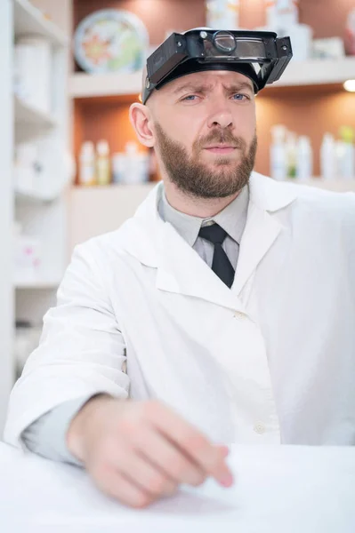 Homme barbu médecin dans un manteau médical et avec une loupe sangle w lumières assises sur le bureau. Equipement optométriste. Cabinet médical. — Photo