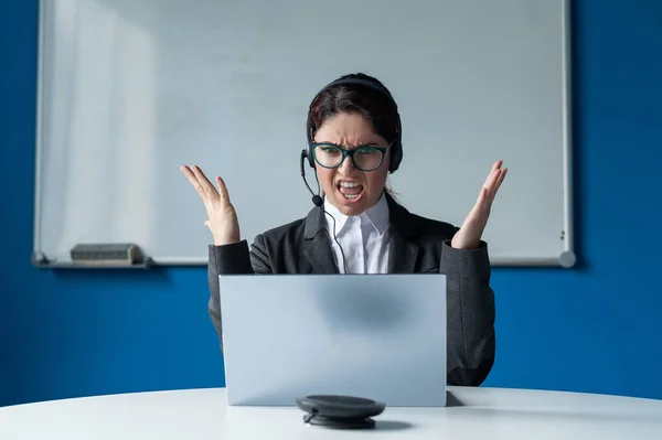 En arg kvinna i ett headset har ett online-samtal på en bärbar dator i ett konferensrum. Kvinnlig chef som skällde ut underordnade för ett videosamtal. Framgångsrika förhandlingar på distans. — Stockfoto
