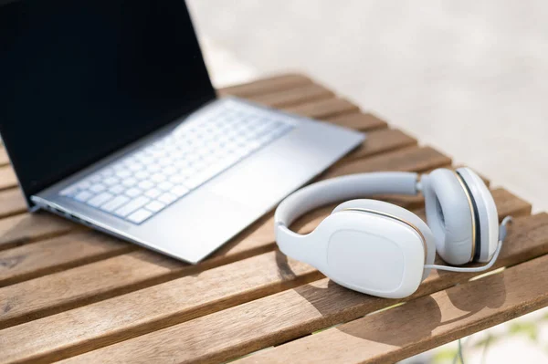 Egy laptop és egy fehér fejhallgató közelsége egy fából készült asztalon egy nyári teraszon. Nincsenek emberek. Kávézó a szabadban. Szabadúszó munkaterület. — Stock Fotó