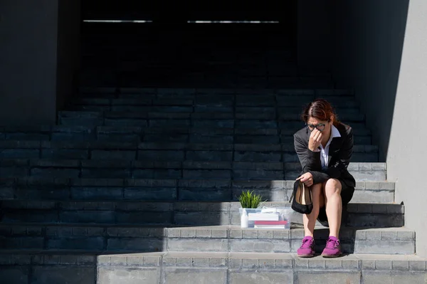 Mujer deprimida vestida con máscara médica es despedida y está sentada en las escaleras con una caja de pertenencias personales. Trabajadora de oficina con traje y zapatillas al aire libre. El desempleo en la crisis económica —  Fotos de Stock