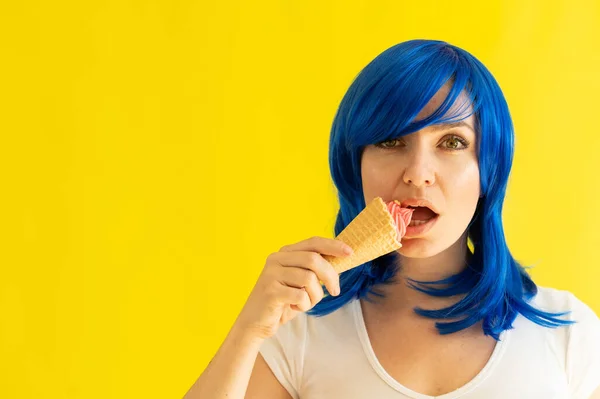 Портрет красивой женщины с голубыми волосами наслаждается поеданием малинового мороженого на желтом фоне. Девушка в парике держит мороженое в вафельной чашке. Летний охлаждающий десерт. — стоковое фото