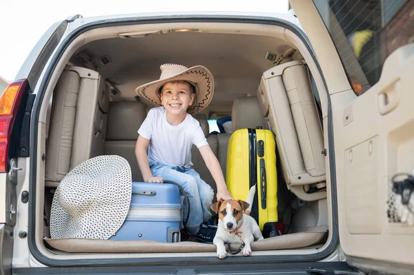 Glücklicher Junge mit Cowboyhut und Welpe Jack Russell Terrier reisen mit dem Auto. Ein Kind und ein lustiger kleiner Hund sitzen im Kofferraum und sind bereit für die Sommerferien. Unabhängiges Reisen. Beste Freunde. — Stockfoto