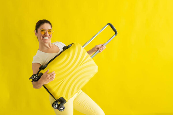 Smiling woman in sunglasses fooling around and holding a suitcase like a guitar on a yellow background. An excited girl in anticipation of a summer vacation trip simulates playing a string instrument.