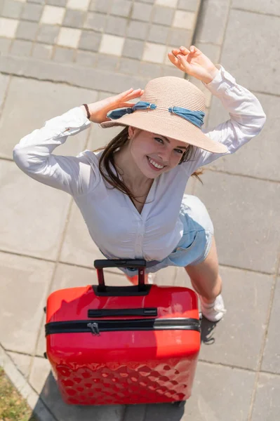 Top uitzicht op mooie jonge Kaukasische vrouw in de zomer hoed poseren met rode koffer buiten. Gelukkig lachend meisje gaat op vakantie met een grote bagage tas. Charmante dame in korte broek op een hete zonnige dag. — Stockfoto