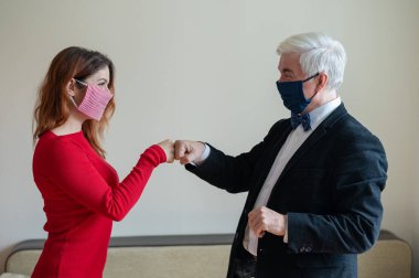 Takım elbiseli olgun gri saçlı bir adam ve kırmızı elbiseli orta yaşlı bir kadın maske takıyor ve yumruk tokuşturuyor. Meslektaşlar sosyal mesafeye saygılıyken selamlaşırlar.