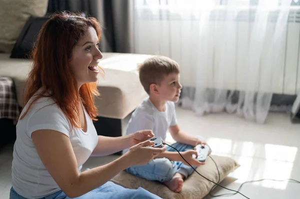Happy woman and son play video games at home. A gambling boy and his mother are playing with the game console joysticks in their hands. A happy family.