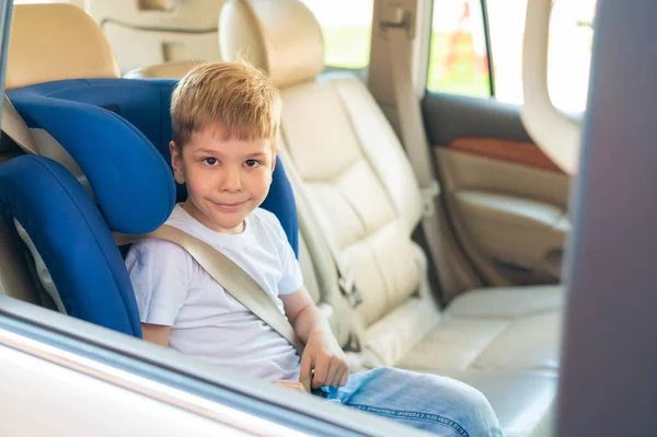 O menino caucasiano senta-se num assento azul de segurança infantil. Viajar com uma criança em um carro com um interior de couro. Retrato de um pré-escolar bonito usando um cinto de segurança e pronto para montar . — Fotografia de Stock