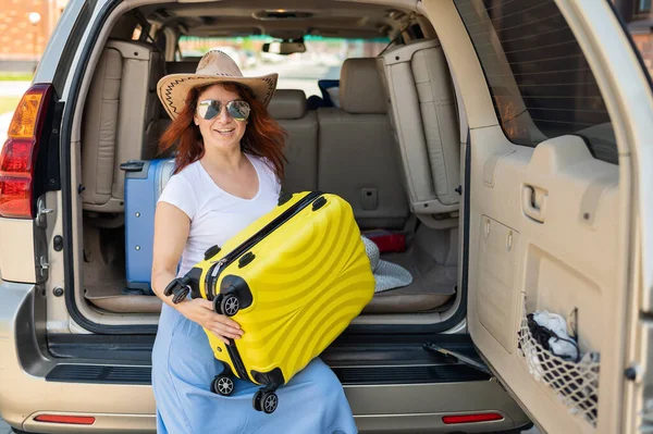 Glückliche rothaarige Frau mit Cowboyhut und Sonnenbrille auf einer Autofahrt durch das Land. Mädchen im Kofferraum mit gelbem Koffer, bereit für den Sommerurlaub. Unabhängiges Reisen. — Stockfoto