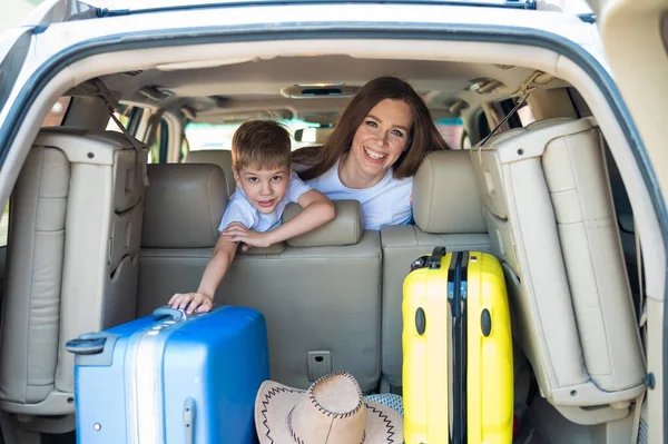 Glückliche Europäerin mit ihrem kleinen Sohn blickt auf den Kofferraum eines Autos mit Koffern. Sommerurlaub für die Familie. Mit einem Kind im Auto unterwegs. Ein Junge mit seiner Mutter auf dem Rücksitz eines Jeeps. — Stockfoto