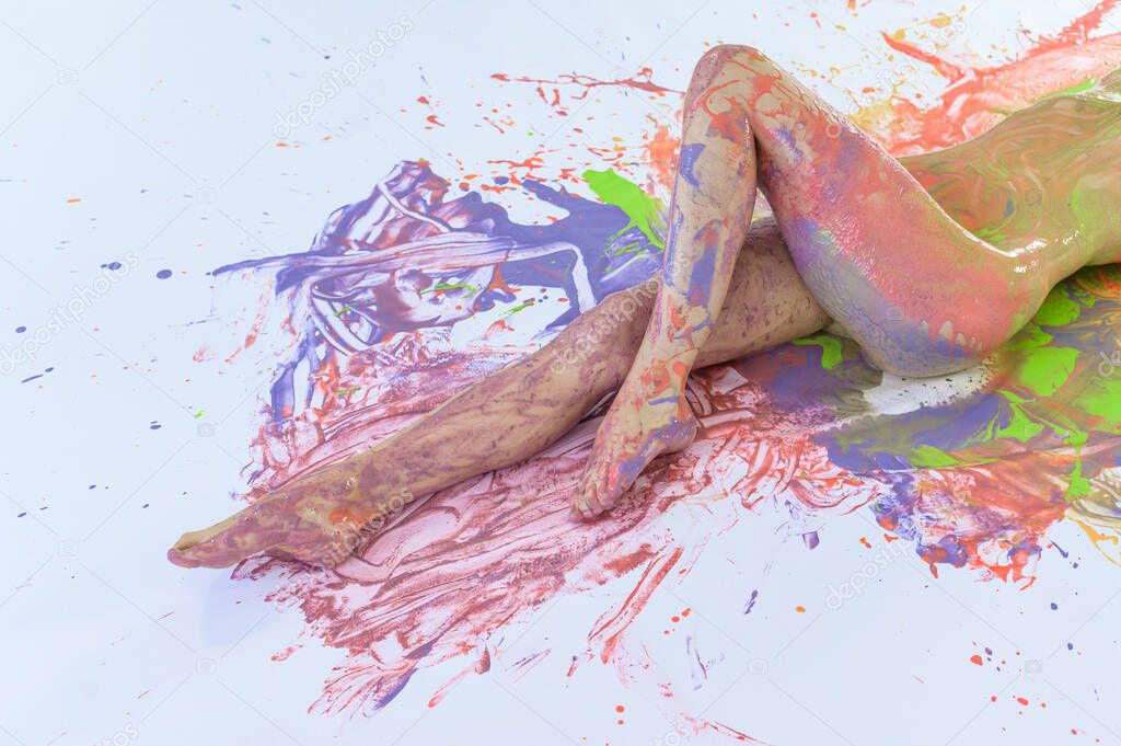 Nude woman in stains of paint on a white floor. View from above. Cropped. Color splashes on the skin. Art body painting.