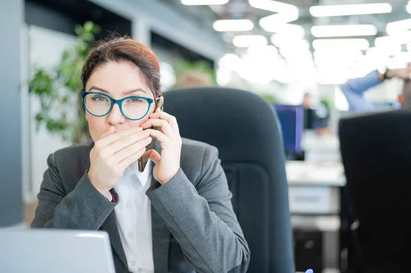 Una mujer se sienta en una oficina en su escritorio y chismorrea por teléfono. Ética corporativa. La empleada de traje cuenta secretos en un smartphone en el trabajo . —  Fotos de Stock