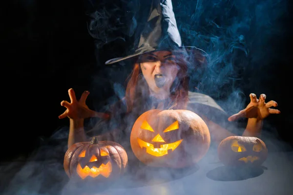 Una bruja siniestra con sombrero evoca sobre una linterna de gato. Personajes tradicionales de Halloween. Niebla mística se arrastra sobre calabazas con caras terribles talladas. Una bruja malvada lanza un hechizo. — Foto de Stock