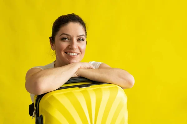 一个带着手提箱的美丽的高加索女人的画像，她梦想着在黄色的背景下度假。一个微笑的女孩在等待旅行时倚在她的行李袋上. — 图库照片