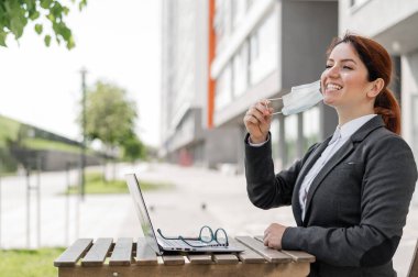 Sosyal mesafeyi korurken dışarıda dizüstü bilgisayarda çalışan tatmin olmuş bir kadın. Gülümseyen kadın ofis çalışanı kafede otururken özgürlüğün tadını çıkarıyor. Coronavirus yenildi. Maskeni çıkar..