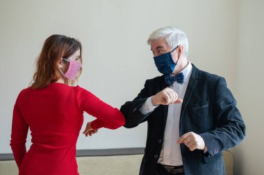 Takım elbiseli olgun, gri saçlı bir adam ve kırmızı elbiseli, tıbbi maske takan kırmızı saçlı bir kadın sosyal mesafeyi koruyup dirseklerini selamlıyorlar. El sıkışmaya ve sarılmaya bir alternatif..