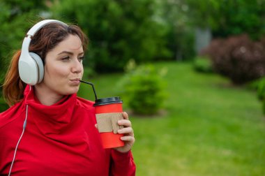 Kızıl saçlı kadın kulaklıkla müzik dinliyor ve parkta pipetle kahve içiyor. Bir kız güneşli bir yaz gününde yürüyüş yapmaktan zevk alır ve bir paket kahve alır. Akıllı telefona bağlı kulaklıklar..