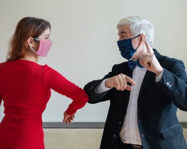 Olgun, gri saçlı bir adam ve kırmızı saçlı, maskeli bir kadın dirseklerini selamlıyor. El sıkışmaya ve sarılmaya bir alternatif. Meslektaşlar aralarındaki mesafeyi koruyorlar. Popüler bir selamlama şekli..