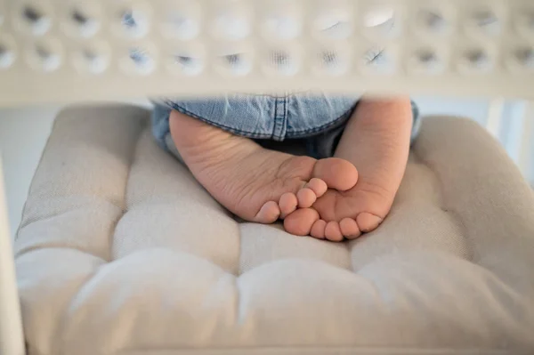 Un chico descalzo se sienta de rodillas en una silla ligera y suave en casa. Los pies desnudos de los niños. Vista trasera . — Foto de Stock