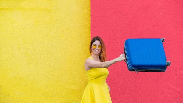 Fröhliche Frau in Peitsche vor gelb-rotem Hintergrund schwenkt freudig einen Koffer in Vorfreude auf die Reise. Schöne Frau mit Sonnenbrille und blauer Tasche in der Hand. Gepäck für den Sommerurlaub. — Stockfoto