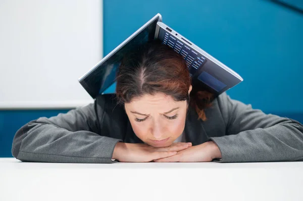 Problemer på jobben. En utslitt kvinne brettet laptopen sin over hodet ved pulten sin. Forretningskvinne i frustrasjon begravd under datamaskinen. Reduksjon og arbeidsledighet. – stockfoto