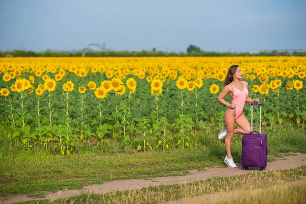 Mulher bonita em um maiô rosa viaja pelo campo com uma mala. Uma menina em um monokini está andando sozinha em um campo de girassóis com um saco grande. Caminhadas. Descanso e distância social . — Fotografia de Stock