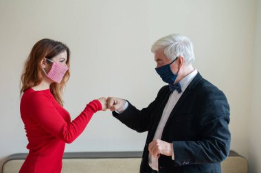 Takım elbiseli olgun gri saçlı bir adam ve kırmızı elbiseli orta yaşlı bir kadın maske takıyor ve yumruk tokuşturuyor. Meslektaşlar sosyal mesafeye saygılıyken selamlaşırlar.
