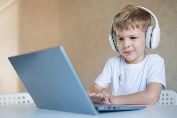 Lindo niño está viendo dibujos animados o una película en un ordenador portátil. Niño caucásico se sienta en una mesa en casa y escucha música en los auriculares. — Foto de Stock