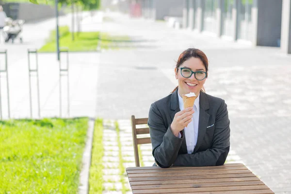 La mujer sonriente disfruta comiendo un cono de helado mientras está sentada en una mesa de madera al aire libre. Retrato de una chica en un traje de negocios para un descanso en un café de la calle en un día caluroso de verano . —  Fotos de Stock