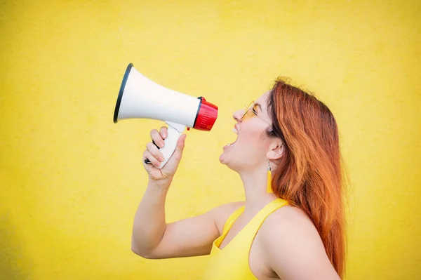 A woman in a dress with glasses and earrings stands in profile on a yellow background and shouts in a megaphone. Portrait of a girl holding a loudspeaker. Lady yells at a sound amplification device.