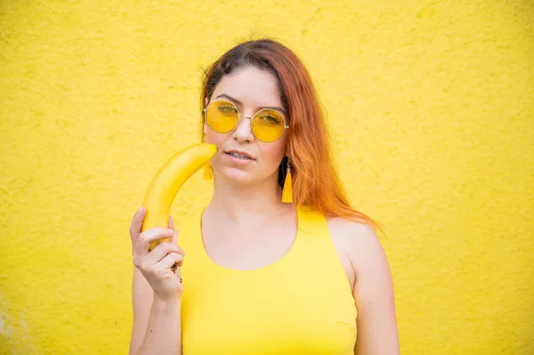 노란 옷을 입고 선글라스를 쓴 아름다운 빨간 머리의 여성의 모습은 감각적 인 입술 근처에 바나나를 들고 있다. 노란 배경에 과일을 곁들인 소녀. — 스톡 사진