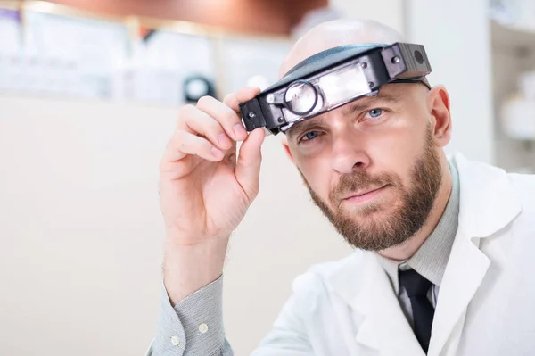 Man skäggig läkare i en medicinsk rock och med en förstorande huvudband w lampor sitter på skrivbordet. Optikerutrustning. Medicinskt kontor. — Stockfoto