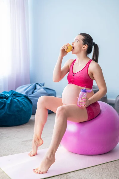 Těhotná žena bez tváře jí po józe jablko. Očekávaná matka kouše ovoce a pije vodu po Pilates. Dívka sedící na fitness míči. — Stock fotografie